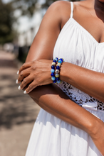 Load image into Gallery viewer, A close-up of a woman wearing a stack of three beaded bracelets on her wrist. The bracelets feature blue, white, and yellow marbled beads, worn over a white sleeveless dress in an outdoor setting.
